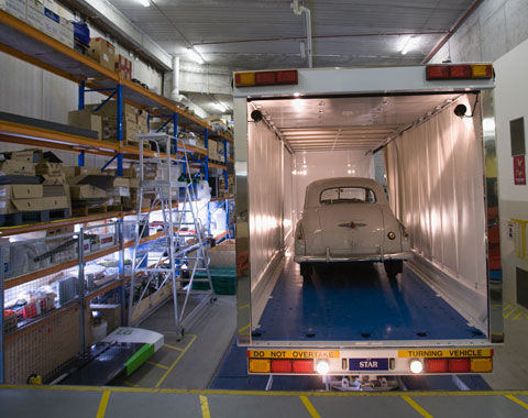 A colour photograph showing an early model Holden car being loaded into a van for transport to a storage facility. The van is at the right side of the photograph, with its rear end closest to the camera. The rear door is open, and inside can be seen the rear of the Holden car. The van is parked in what appears to be a loading bay; large shelves at the left of the photograph hold a variety of objects, some of which are in boxes. On the concrete floor of the loading bay can be seen yellow stripes that appear to mark out safety zones.
