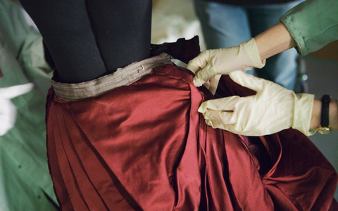 A close up of two glove-encased hands emerging from the right side of the image holding ruby red fabric with a satiny finish. In the background and out of focus, the legs of another person are visible.