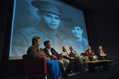 Six members of the forum panel seated on stage with a large image of a young soldier and a woman on a screen behind them.