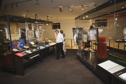 Two long display cases frame either side of this image. The clear cases contain many different objects. A man and a woman stand looking into a case at the left rear.