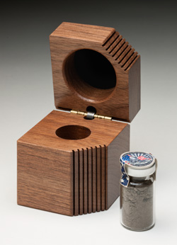 A pentagon-shaped timber box, etched with vertical grooves at the centre. The hinged lid is open and shows a circular hole at centre. A small glass container with a silver lid sits beside the box. It contains soil and has a seal at top with an eagle and the text 'SEPTEMBER 11, 2001'.