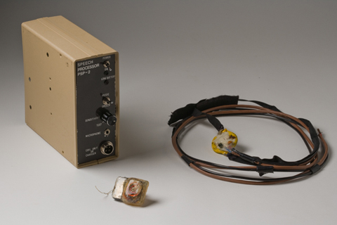 Small bionic ear implant, with coiled cables inside, pictured with a box marked 'Speech Processor PSP-2' and a small receiver unit attached to coiled cables and a velco headstrap.