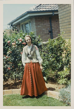 Colour photograph showing a woman dressed in Latvian national costume, consisting of a red skirt, beige waistcoat, white shirt and cloth cap. The woman stands in a suburban garden.