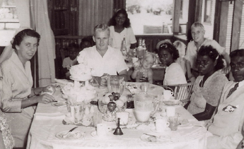 Group of adults sitting at a large tble in the foreground, with children seated at a smaller table behind.