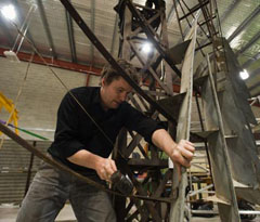 A man stands behind a metal windmill frame. He holds rubber mallet in one hand, and a steel fan in the other.