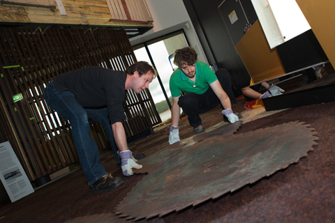 A colour photograph of two men laying metal objects on a floor.