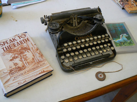 An old-fashioned typewriter on a desk. The typewriter has round keys and the word 'Corona' on the front of the mechanism casing. A piece of string is tied to the front of the typewriter; a disc with a hole in it is attached to the other end of the string. Two books are next to the typewriter. The book at left is called Speak To The Earth while the book at right is called The Silver Brumby.