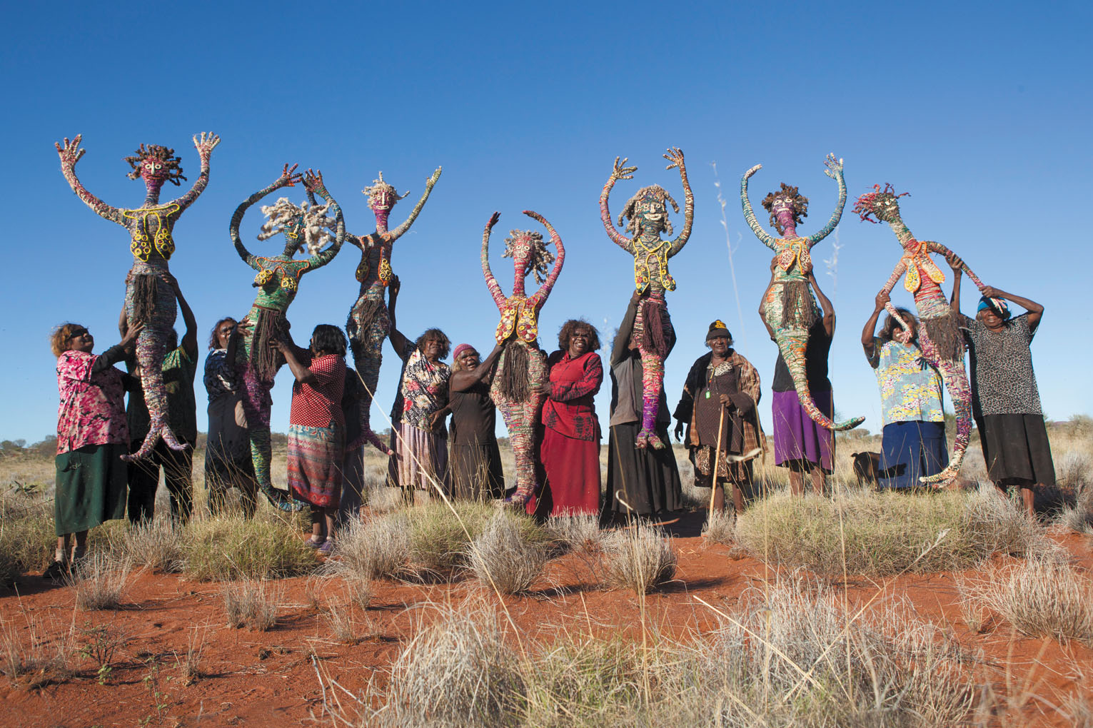 Group of women in a desert holding large grass figures above their heads.