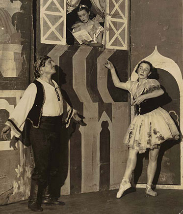 Three performers on stage. Two of them, a man and a woman in traditional folk costume, strike balletic poses while another performer reads a book in a window above their heads