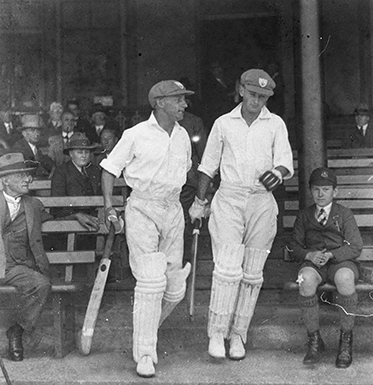 Two white-clad batsmen emerge from the stands with spectators on wooden benches on either side.