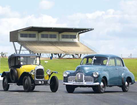 A 1923 5CV Citroën car, with yellow bodywork and black roof, and the blue-grey 1946 Holden Prototype No. 1 sedan at the Oran Park raceway. A grandstand is visible in the rear.