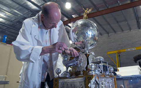 Medium distance shot of a conservator working on a silver trophy. The conservator, wearing a white lab coat, is on the left side of the image facing the camera but with his face turned towards the object. The object, on the right is a large silver globe with Australia in relief and clearly visible. A golden figurine perches on the top of the globe and various figures of animals are at the base of the trophy.