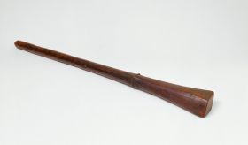 Club made of brown wood with a handle that is slightly rhomboid shaped at one end and pointed at the other. Both ends separated by a bulged ring.