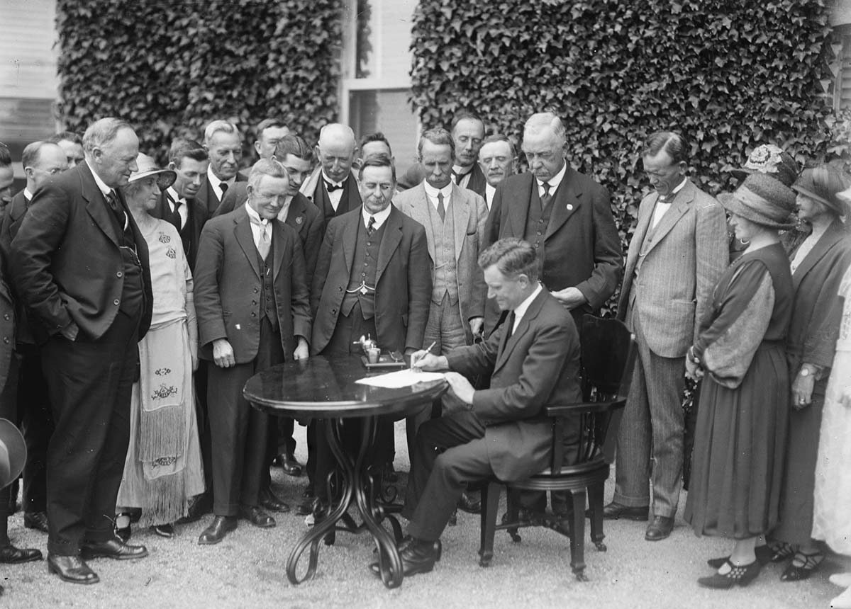 Black and white photo of a group of people watching a seated man at a table signing a document.