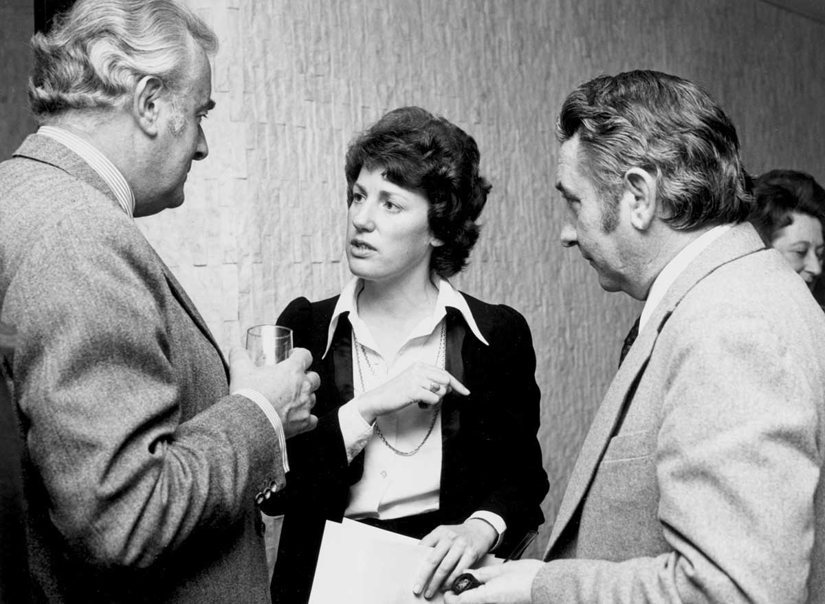 Interior photo. Woman in her thirties is speaking while Whitlam and Oswin listen