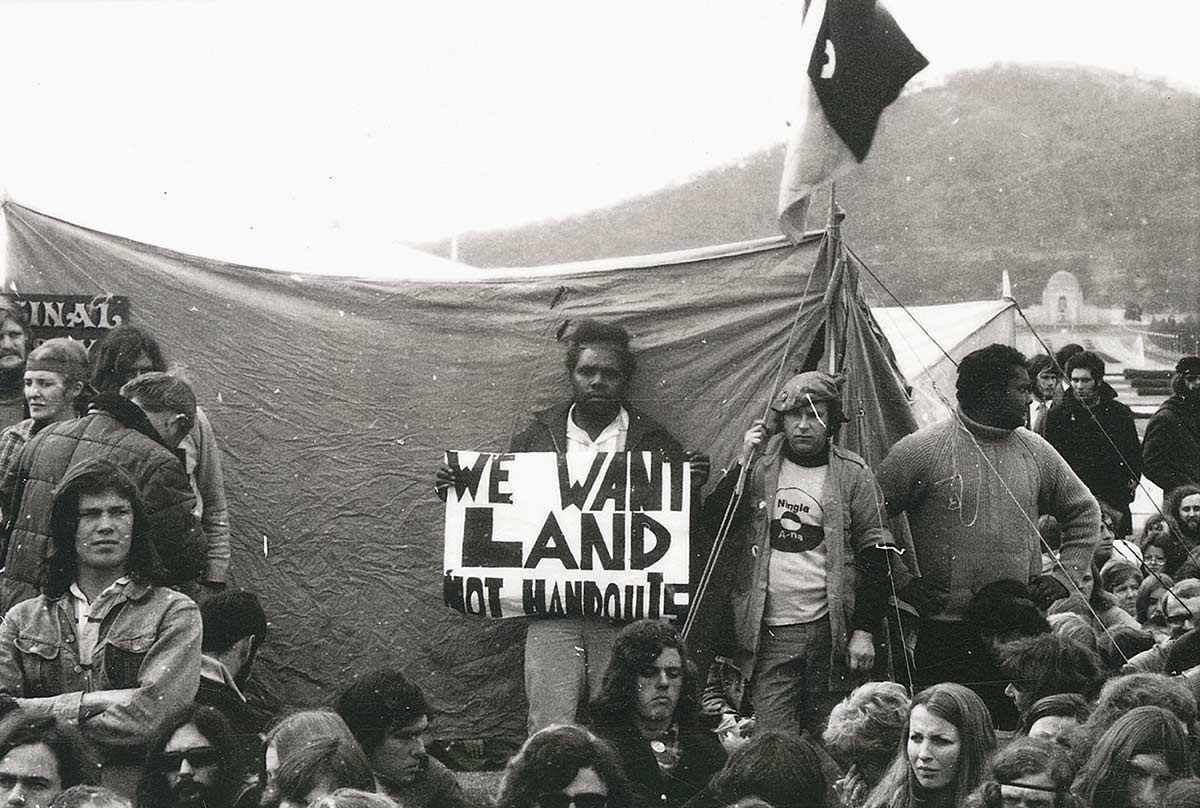Aboriginal man standing in front of tent holding up sign saying ‘We want land not handouts’. He is surrounded by a dozen or so other protesters.