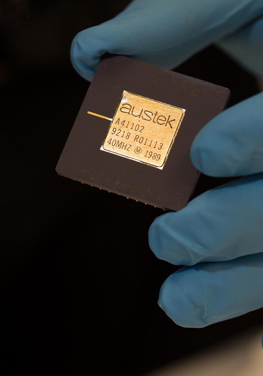 a hand wearing plastic glove holds coin-sized black square with gold-coloured chip at its centre - click to view larger image