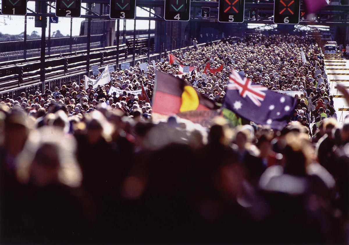 A vast crowd of people cover the roadway of the Bridge. People are carrying the First Nations and Australian flags.