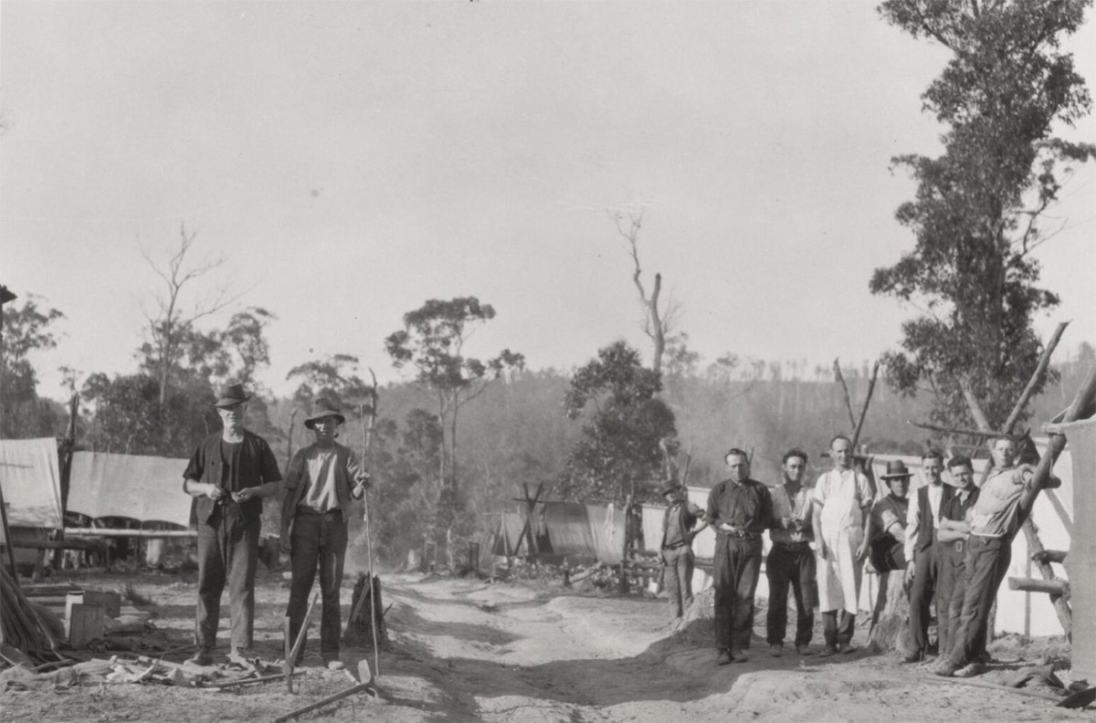 A group of 12 or so men in work clothes pose for the camera. They are in the bush on either side of a dirt track and surrounded by what appear to be tents. It’s possible they’re involved in land clearing.