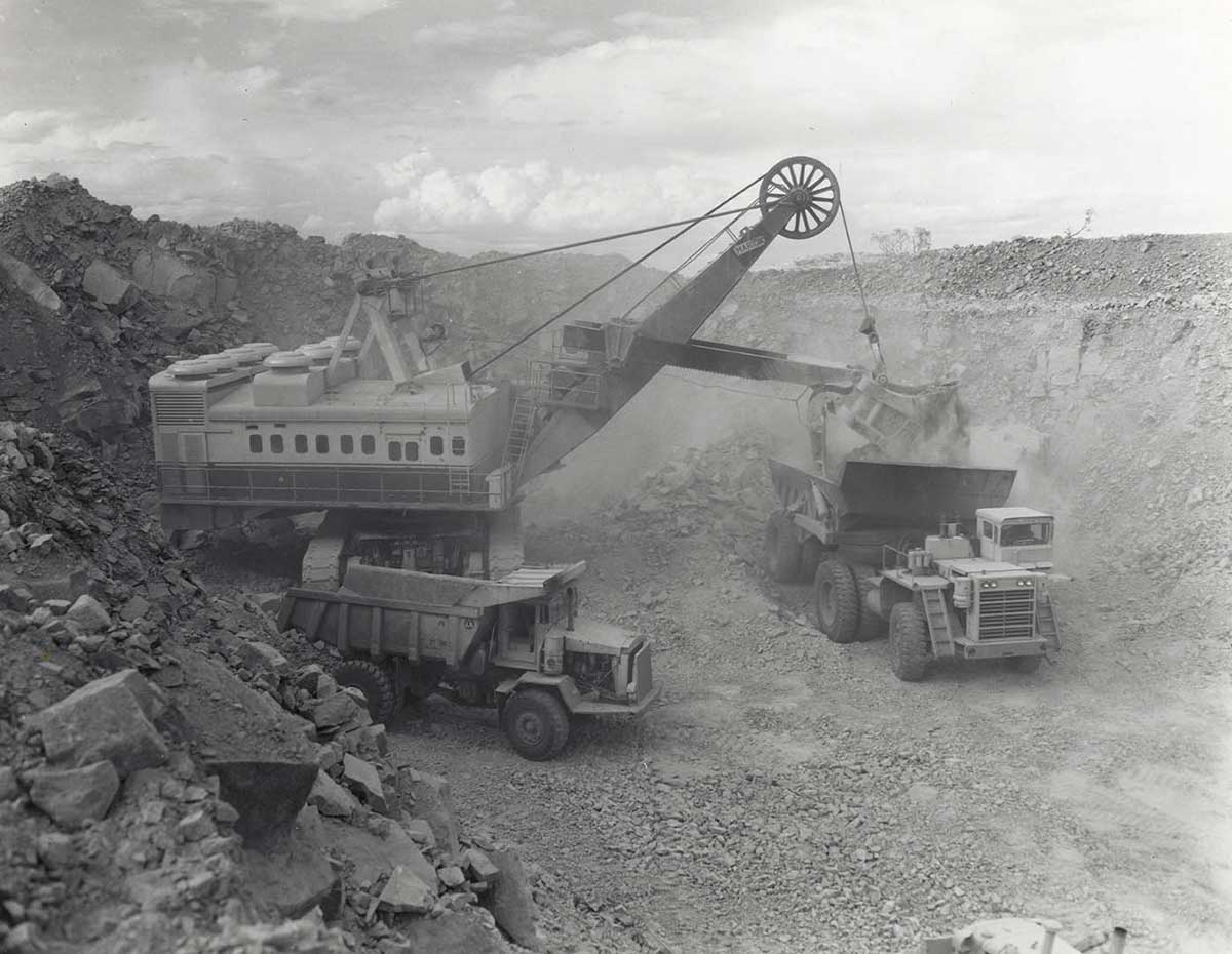 Black and white photo of large power shovel dumping ore into one of two large mining trucks.