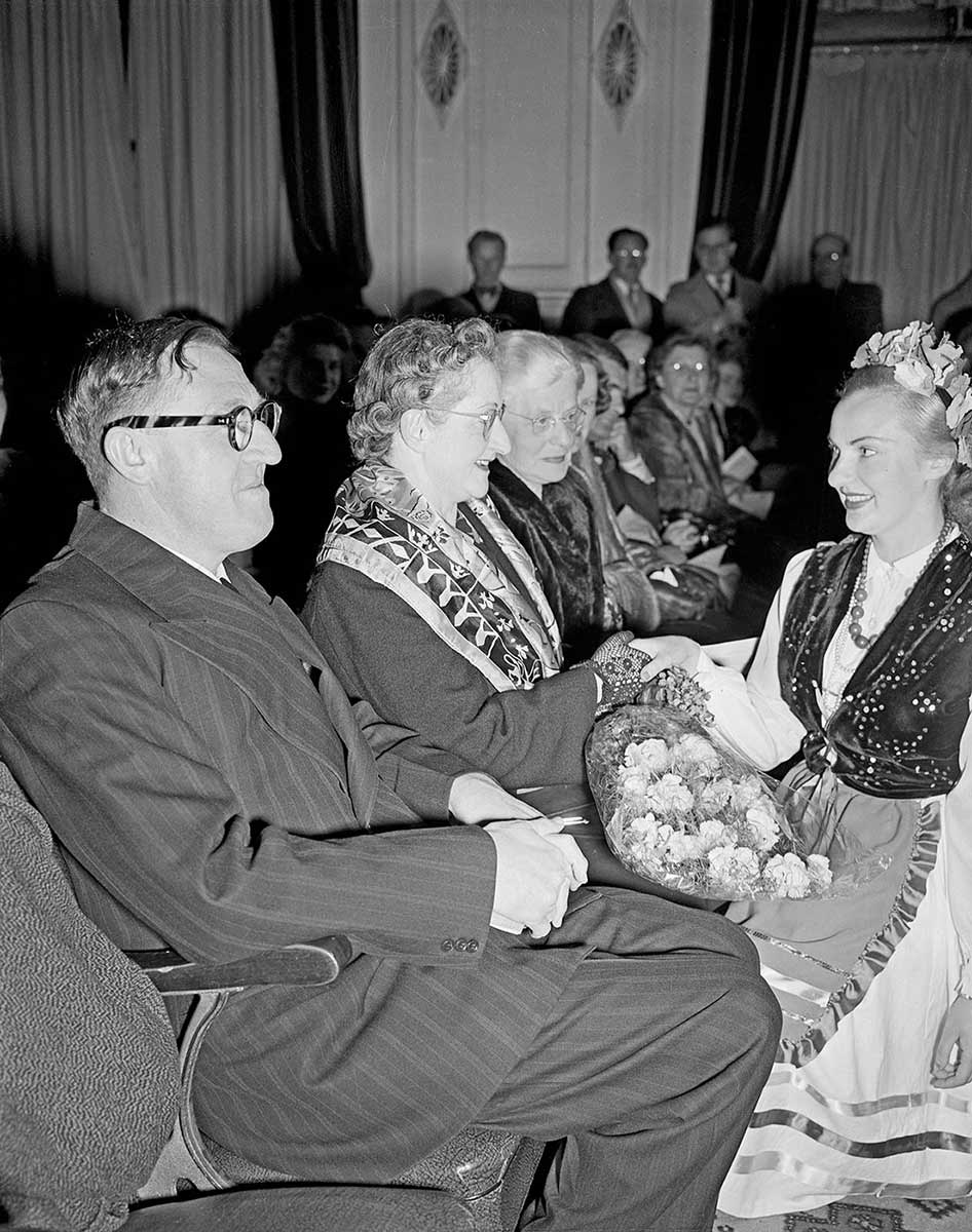 The Calwells seated with a woman in traditional Lithuanian dress shaking hands with Mrs Calwell. - click to view larger image