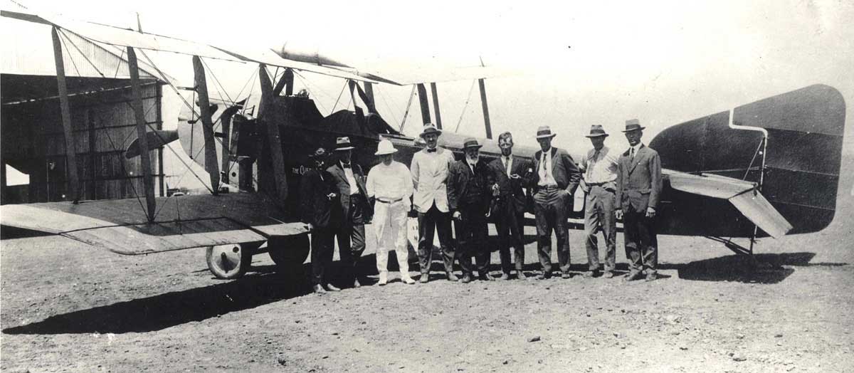 Biplane with nine men standing, posing for the camera, by its fuselage.