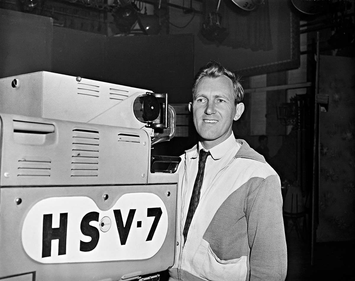Smiling camera man behind large camera with HSV 7 marked on the side. Taken inside a TV studio.