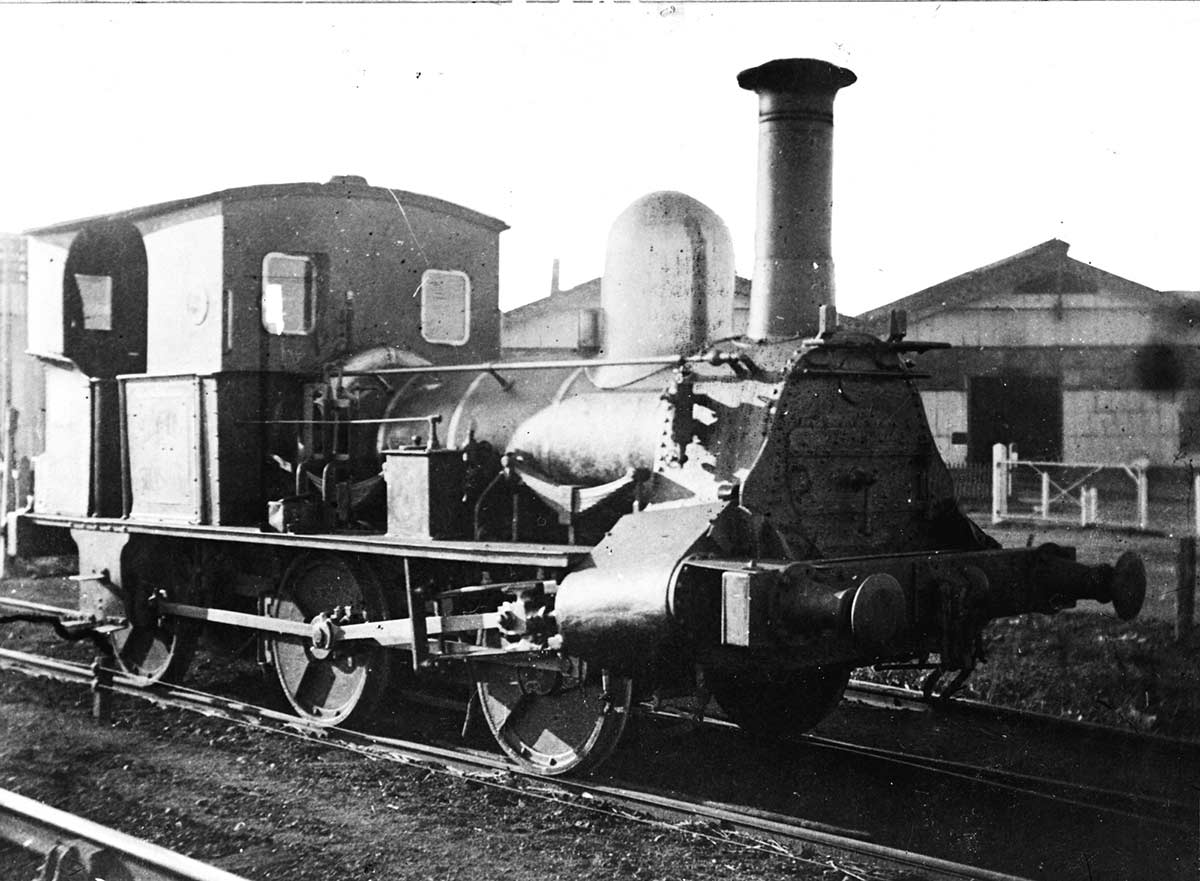 Slightly scratched photo of an early steam engine with railway sheds in the background.