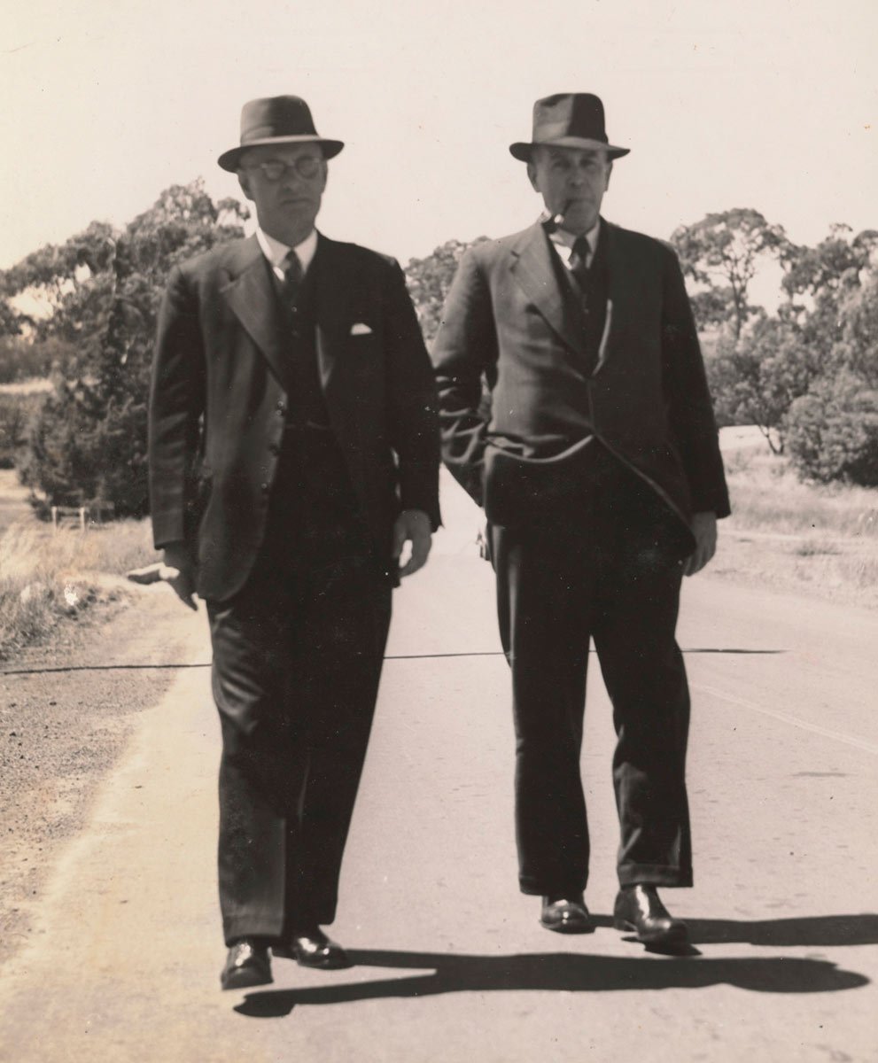 two men in suits and hats walk side by side outside.