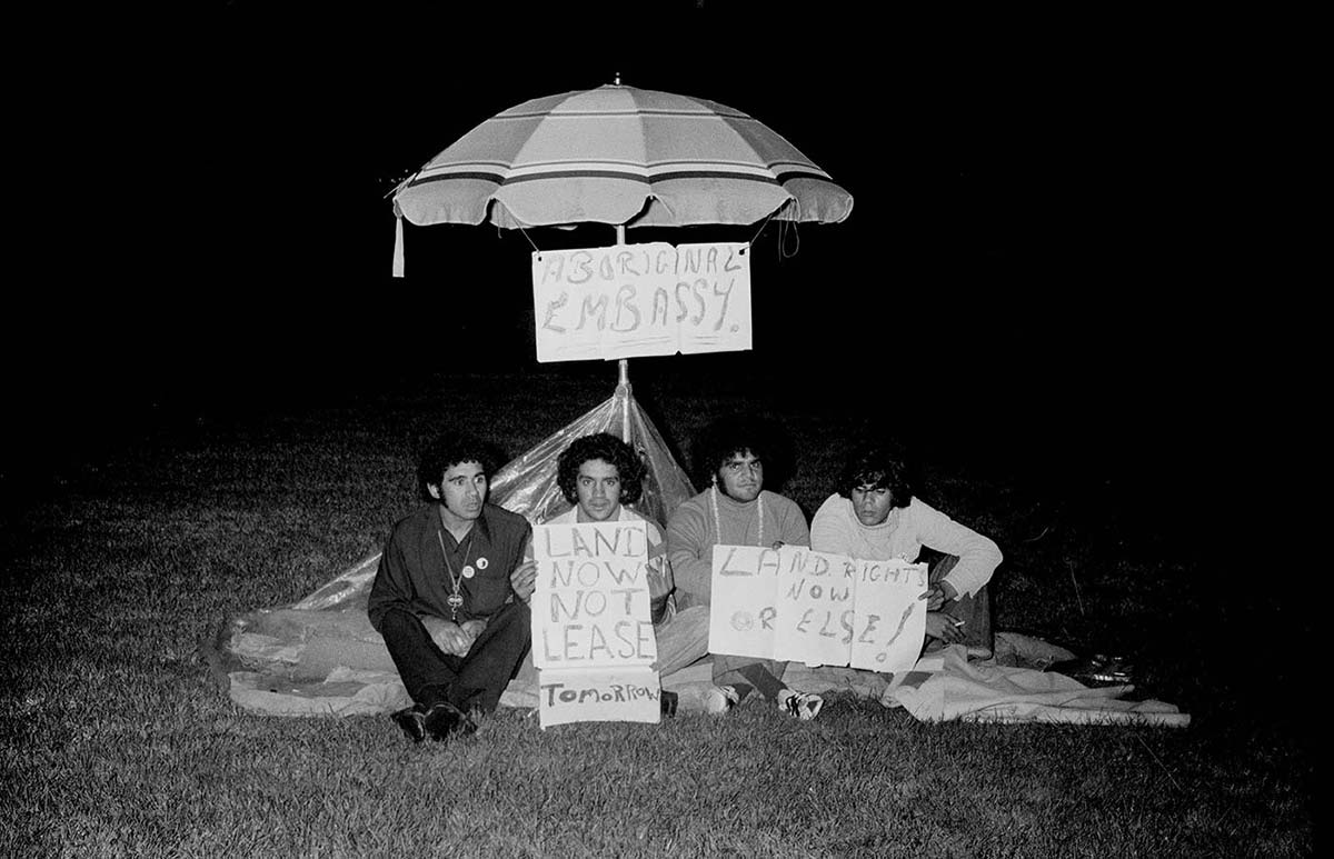 Four young men sit in front of a beach umbrella at night holding up signs.