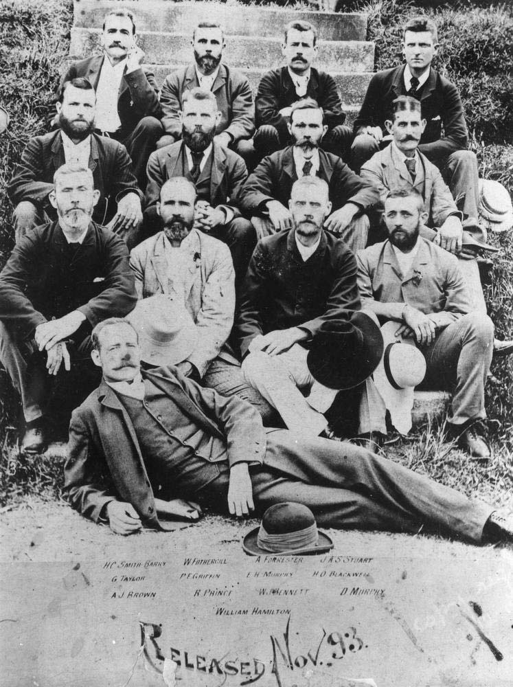 A group of men sitting on a set of stairs. There are four men sitting on each of the first three steps and a single man lying on the ground at the front of them. - click to view larger image