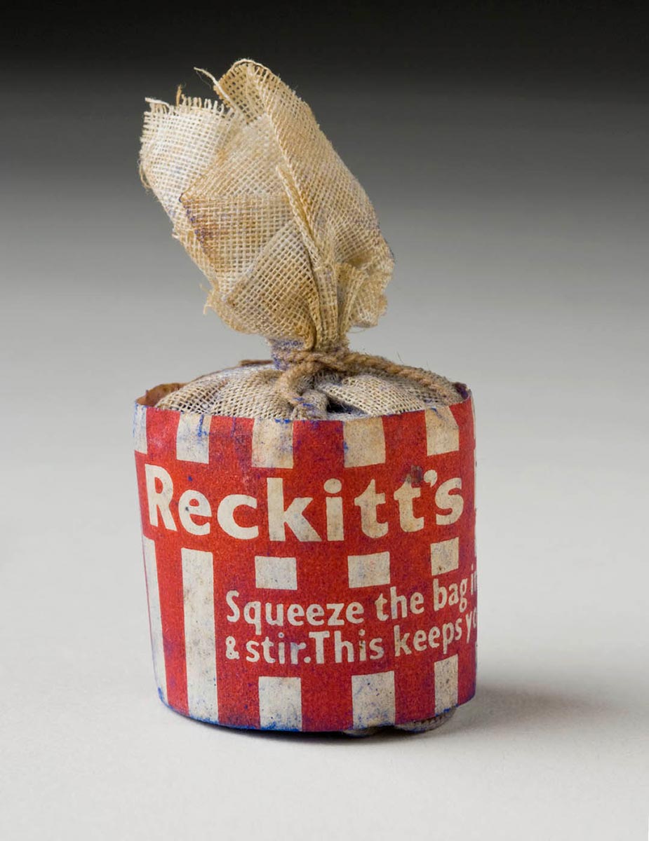 Two small off-white cylindrical cloth bags, tied at the top with string. Red and white striped paper is wrapped around each bag, with the text 'Reckitt's. ASqueeze the bag & stir. This keeps ...' Blue powder is visible on both bags.