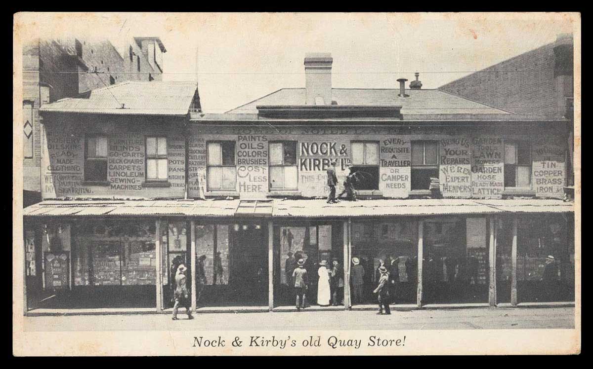 Promotional postcard featuring a black and white photograph of a shopfront with 'Nock and Kirby Ltd' painted on the front. The names of store products are painted on the exterior walls of the second level, and there are a number of people gathered at the entrance. - click to view larger image