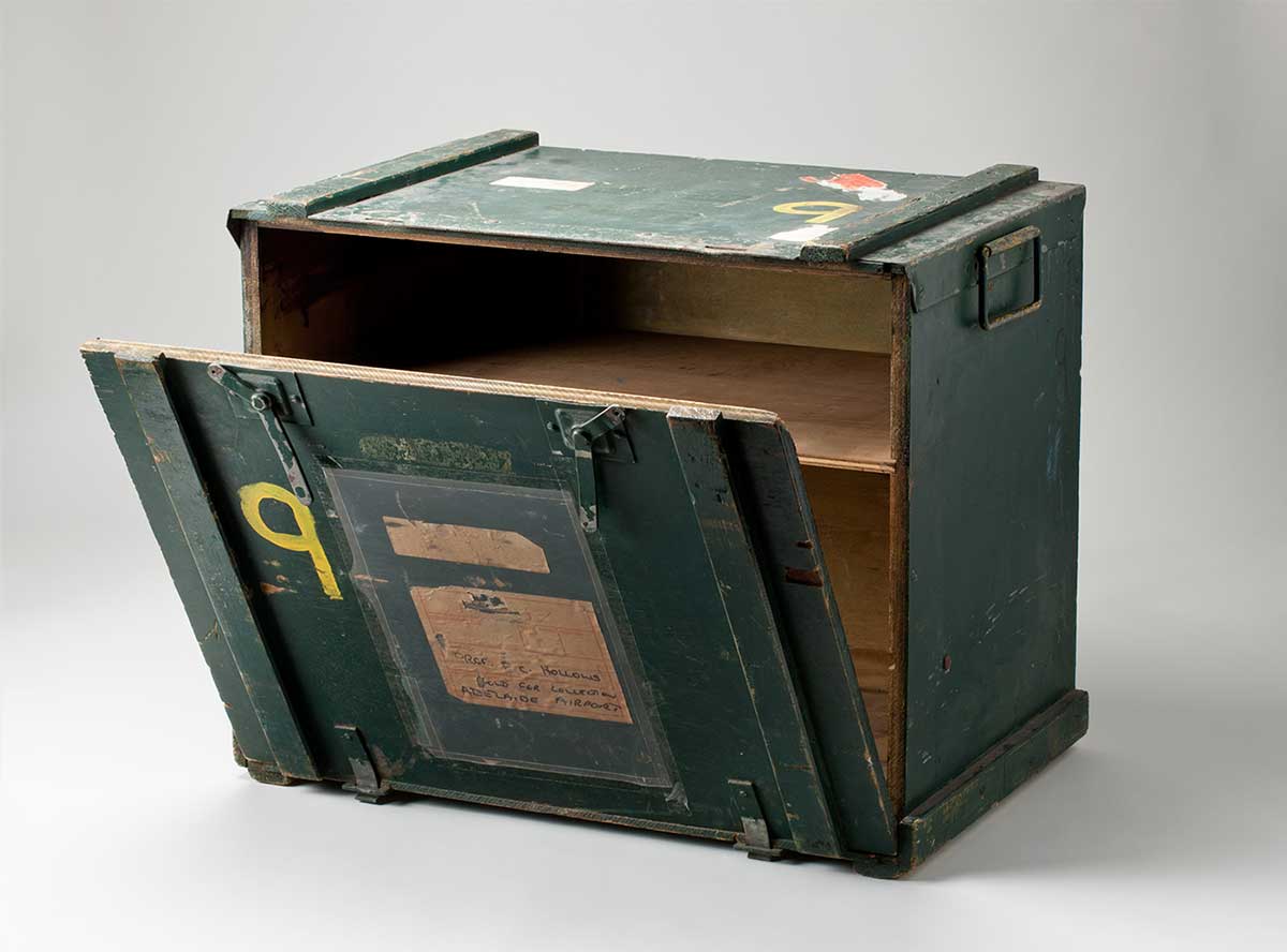 A green coloured wood and metal rectangular storage box with remnant stickers on the top and front. Text on the address label reads 'PROF. F.C. HOLLOWS / HOLD FOR COLLECTION / ADELAIDE AIRPORT'. The number '9' is painted on the front and the lid in yellow. - click to view larger image
