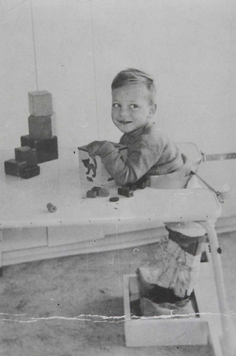 Black and white photo of a young boy at a table. He wears callipers on his legs and is playing with a box and blocks. - click to view larger image