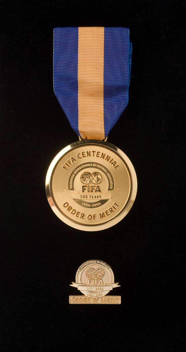 A gold metal disc with a central logo and the text 'INTERNATIONALE DE FOOTBALL ASSOCIATION / FIFA / 100 YEARS / 1904-2004'. The medal hands from a blue and yellow vertically striped ribbon. A small circullar gold finished lapel pin sits below the main medal. Both appear on a black backdrop. - click to view larger image