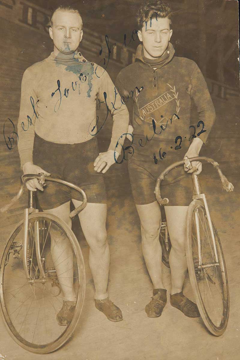 Black and white photograph of two men standing beside their bicycles. - click to view larger image