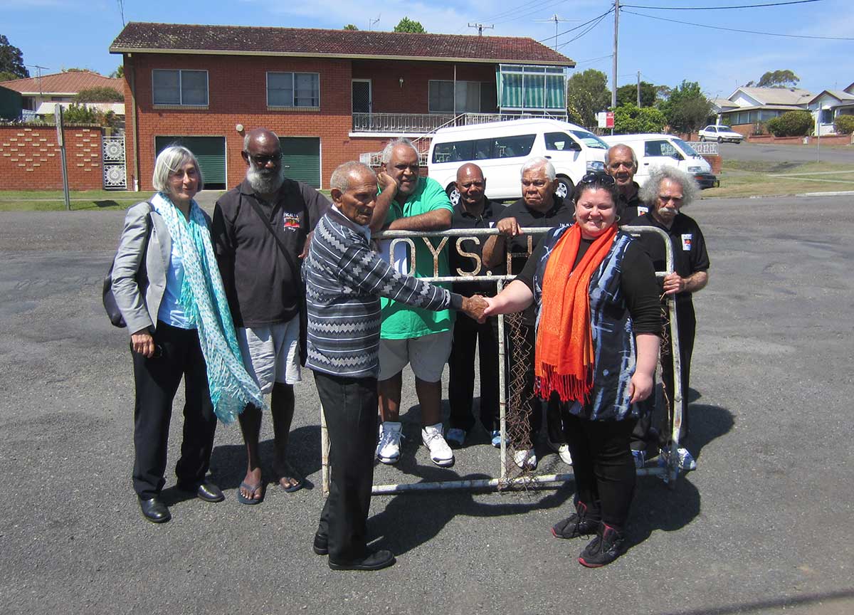 A group of two women and seven men standing in a street. Several of the men lean on a metal gate. A house and two small buses are in the background.