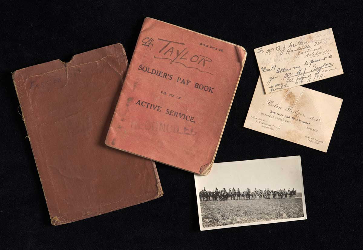 A booklet titled 'Soldiers' Pay Book for use on Active Service' that is stamped 'RECONCILED' and has handwritten on the front 'CPL. TAYLOR.' The book is protected by a brown slipcase with torn edges with 'FARRIER SGT. TAYLOR / 199' in ink on the front. Inserted into the book was a black and white photograph of a line of mounted Light Horsemen. The photograph has 'Empty saddles / after the battle of Gaza' handwritten in pencil on the reverse. There are also two cards with the pay book. One card has finger print smudges on it and reads 'Colin Rodger, M.M / Jeweller and Watchmaker'. The second card reads 'To Mr B.J. Miller ... / 'Bert! allow me to present to you Mr A. J. Taylor a very old friend of mine from the A.L.H' The other side of the card reads 'From / 68 sun / S.S.M.W.G Miller / D.M.C. rest camp'. - click to view larger image