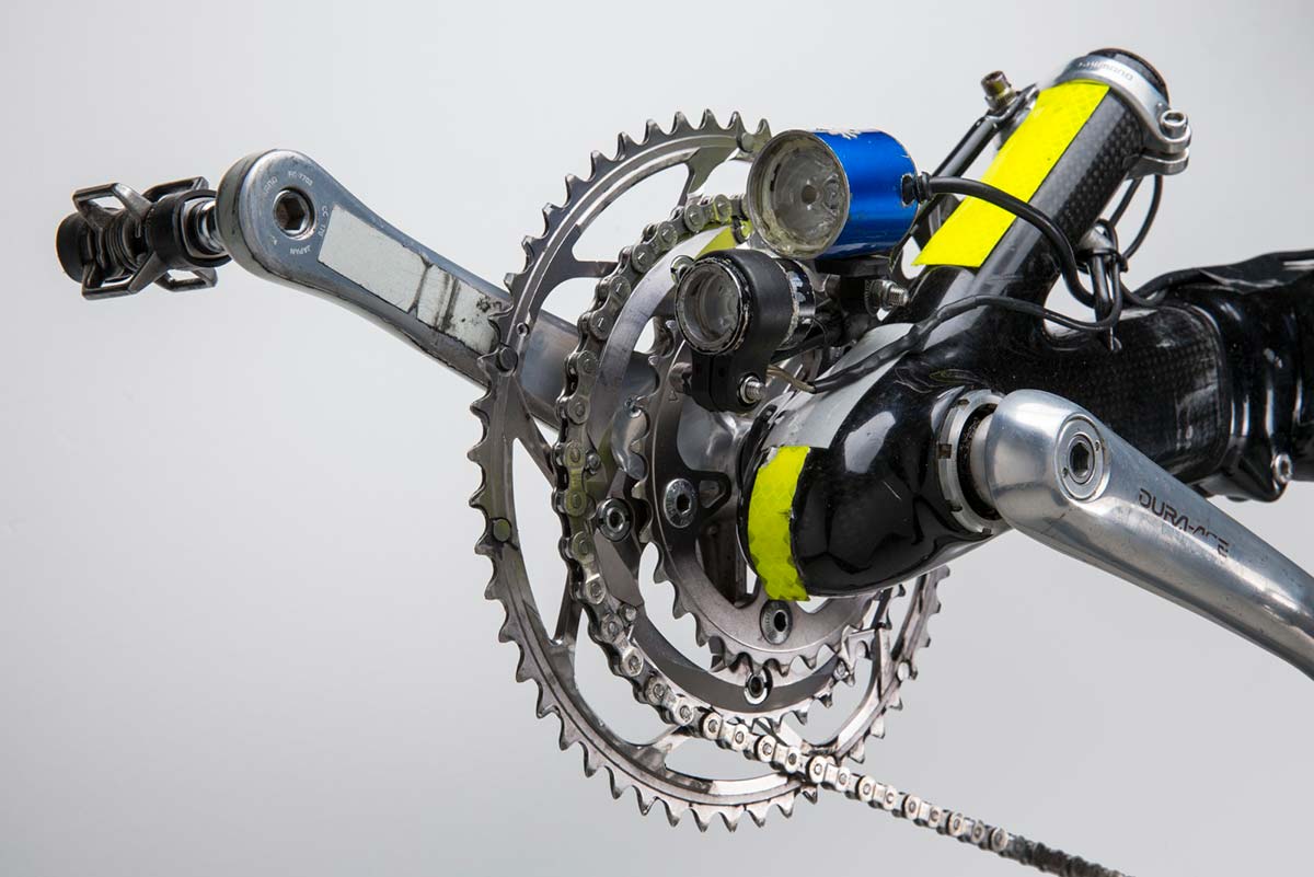 Colour photo showing a closeup view of a bike's crank set, with two small lights mounted on the frame, and two small yellow reflective strips. - click to view larger image