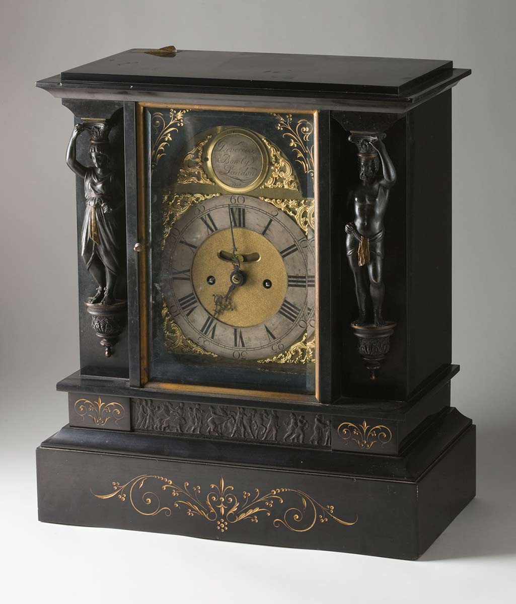 A mantelpiece clock, showing the face and one of the sides. The clock is made from dark material and has carved decorations. - click to view larger image
