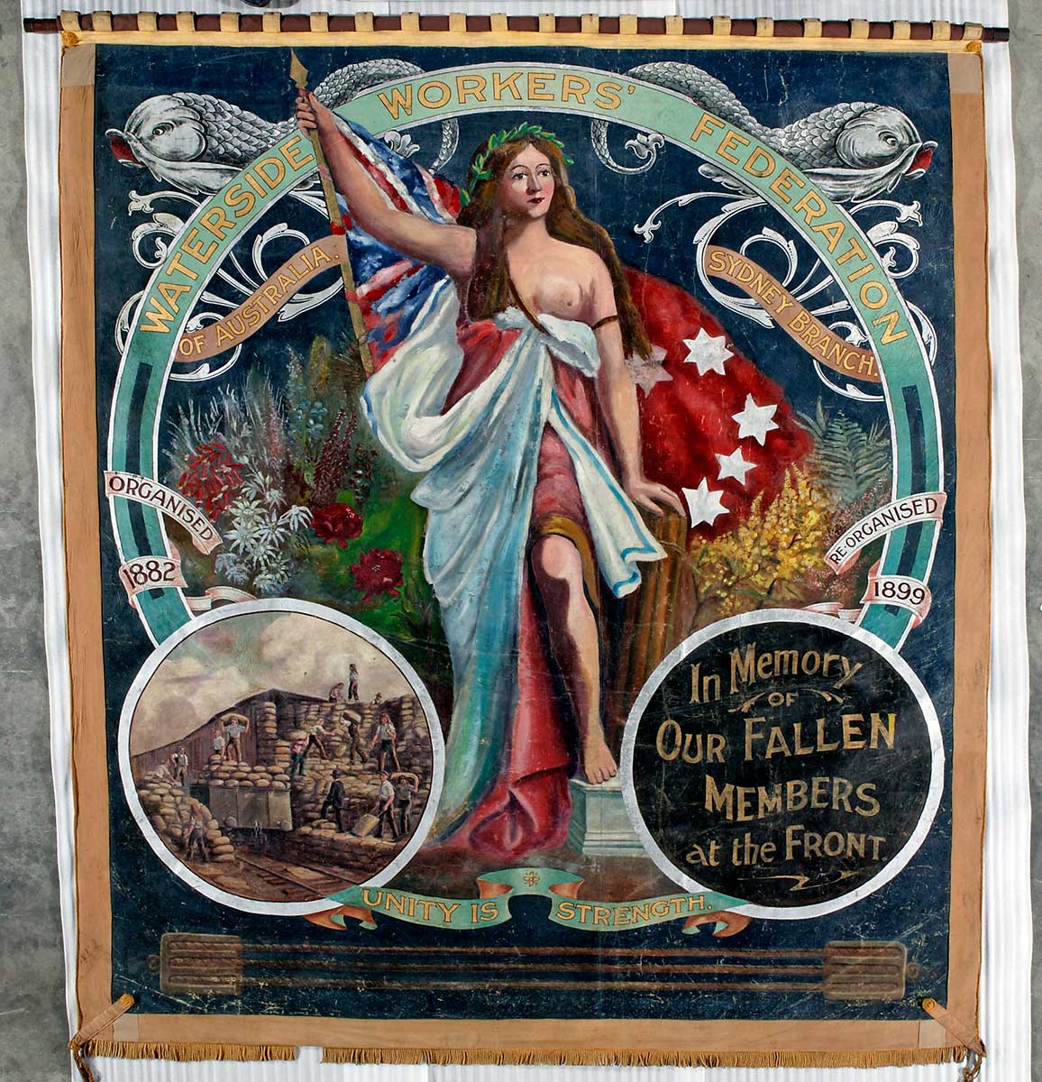 Large, almost square shaped banner painted in oil colours and showing a central image of a woman wearing loose flowing robes. She holds the red ensign and stands among native flowers. The text at the top reads 'Waterside Workers' Federation of Australia, Sydney branch'. An inset image at the bottom left shows workers handling numerous sacks and an inset at the bottom right reads 'In Memory of OUR FALLEN MEMBERS at the Front'. The motto 'Unity is Strength' joins the two insets. - click to view larger image