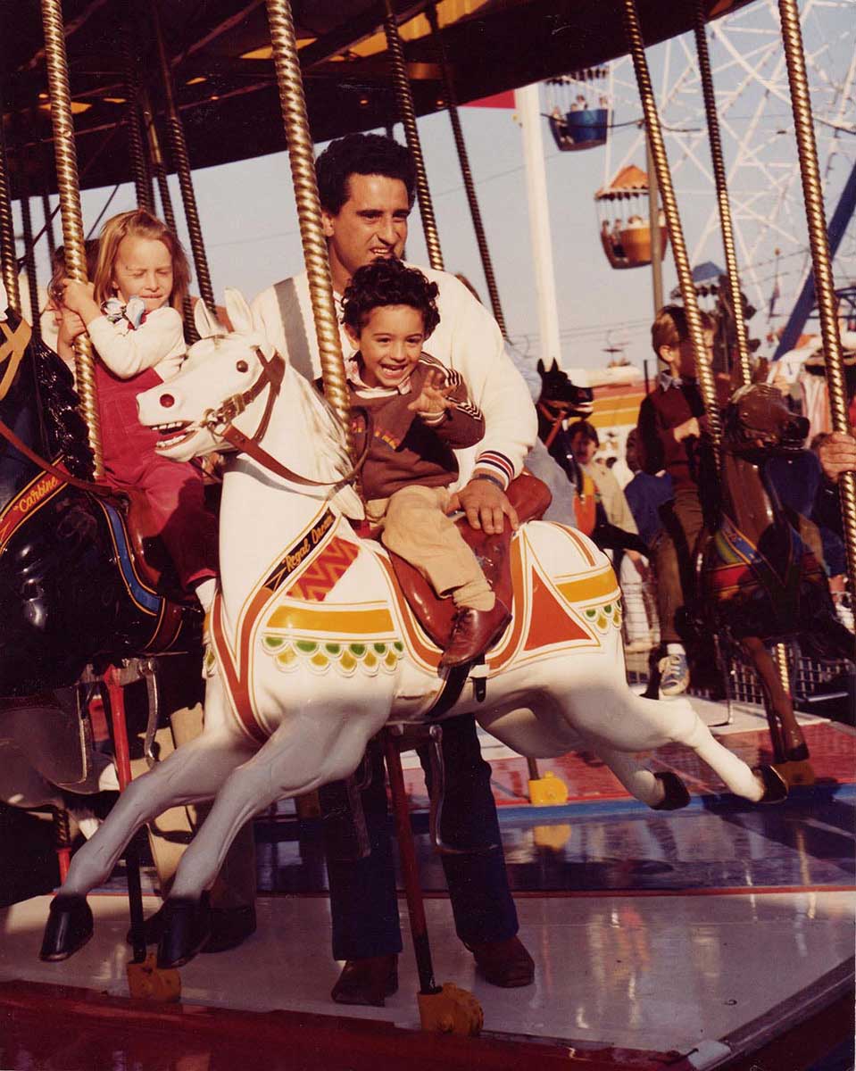 An image of children on a carousel ride.  Featured in the foreground is small boy on a carousel horse with a man standing behind him. - click to view larger image