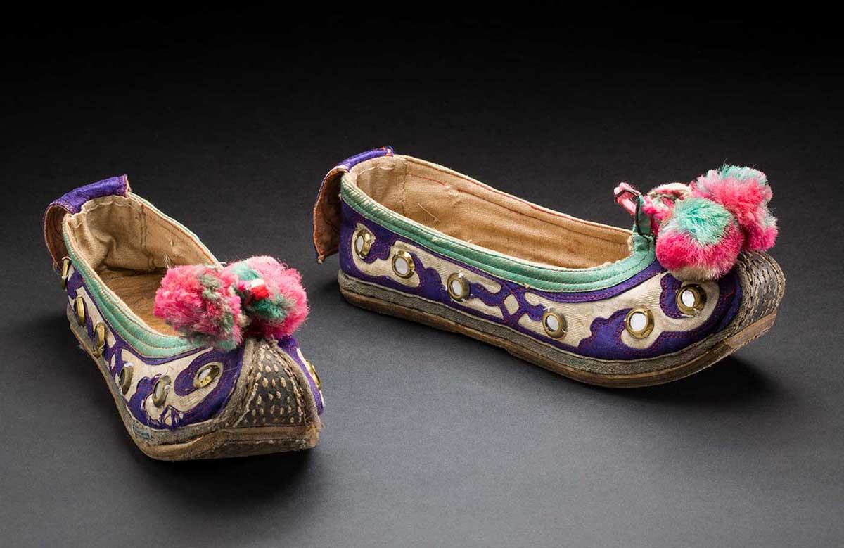 Colour photograph of two embroidered slipper-style shoes with upturned toes and pink and green pompoms at the end. - click to view larger image