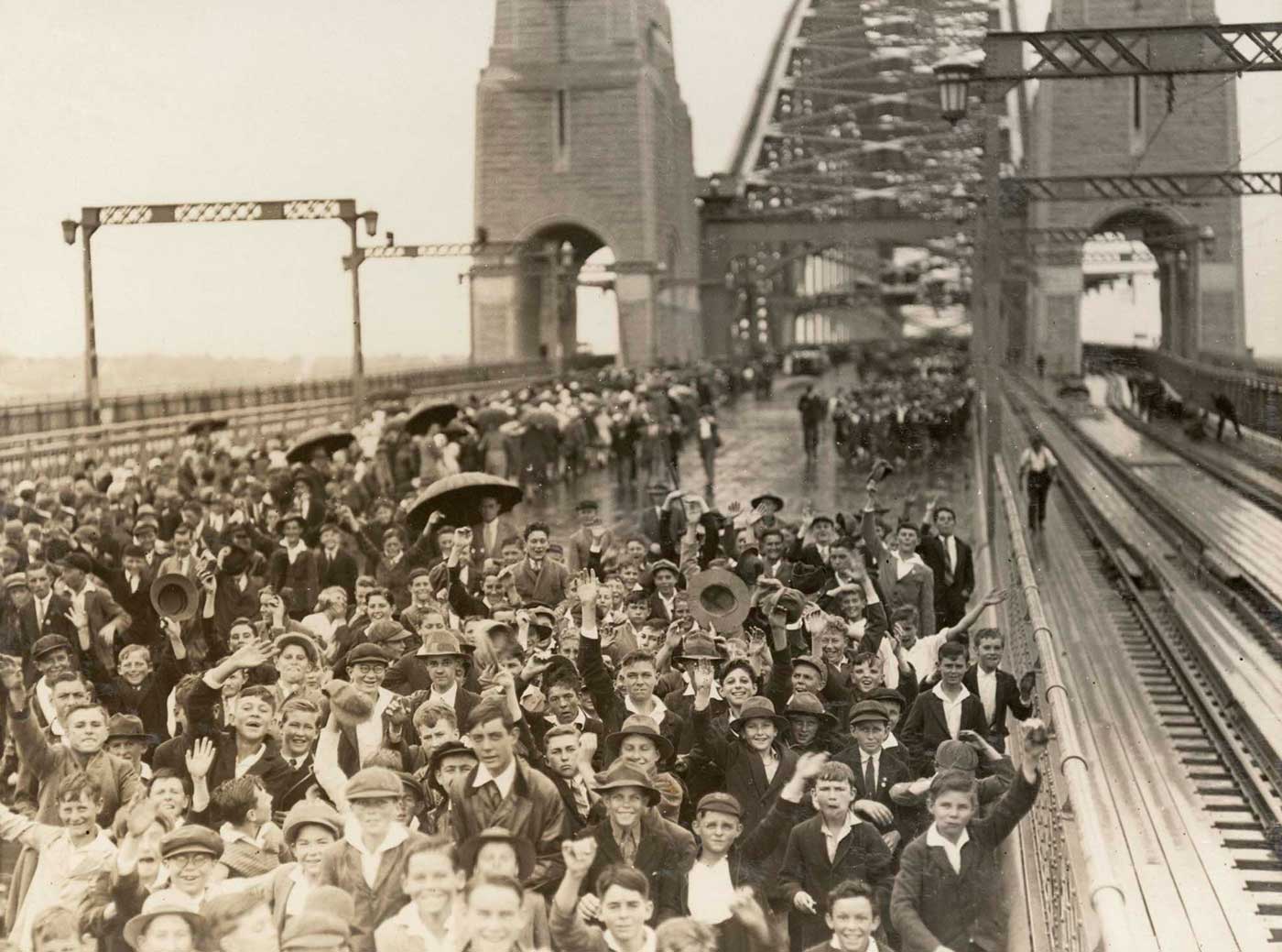 Black and white photo of a large crowd of schoolchildren crossing the Sydney Harbour Bridge. There is no traffic on the bridge. - click to view larger image