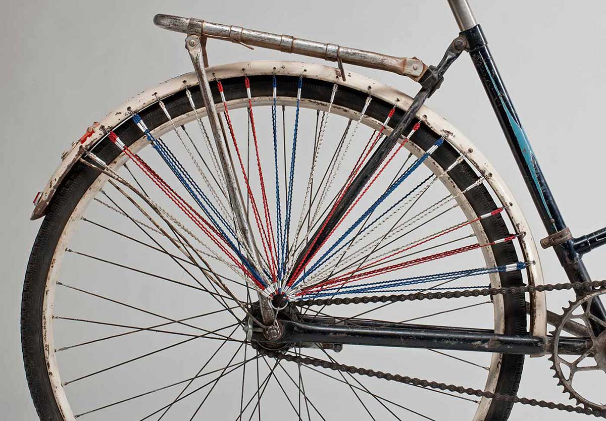 The rear wheel has lengths of red, white and blue fine cord attached through the mudguard and gathering just above the centre of the wheel; on both sides of the rear wheel.  - click to view larger image