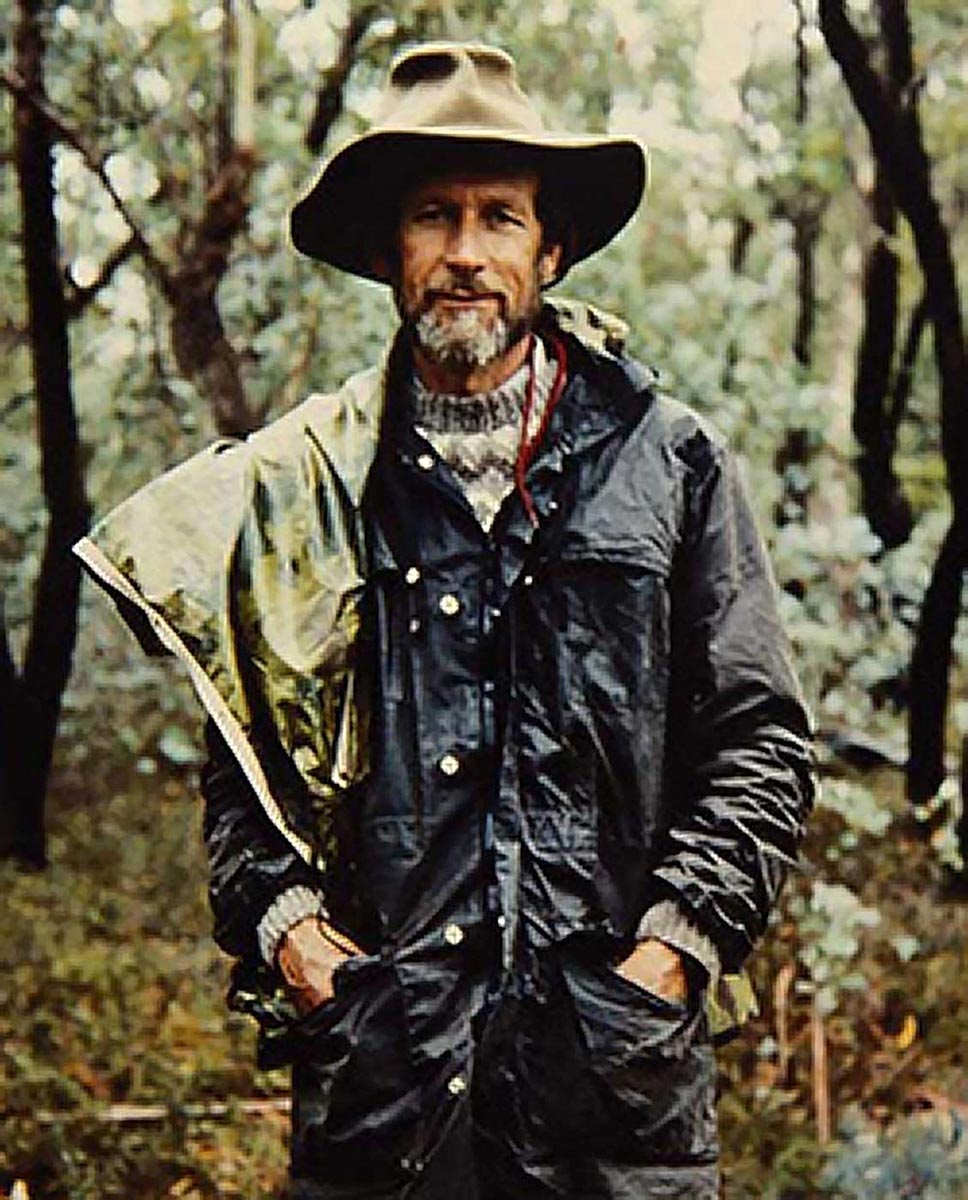 Portrait of a man standing in a forest wearing a hat and waterproof jacket.