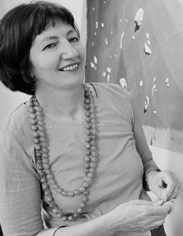 A black and white portrait photo of a woman. Part of an embroidery artwork is hanging on the wall behind her.