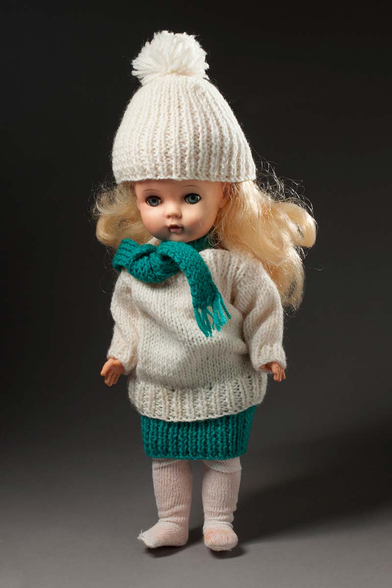 A doll with blonde hair wearing a cream coloured knitted beanie and jumper, a green coloured knitted scarf and skirt and cream stockings. - click to view larger image