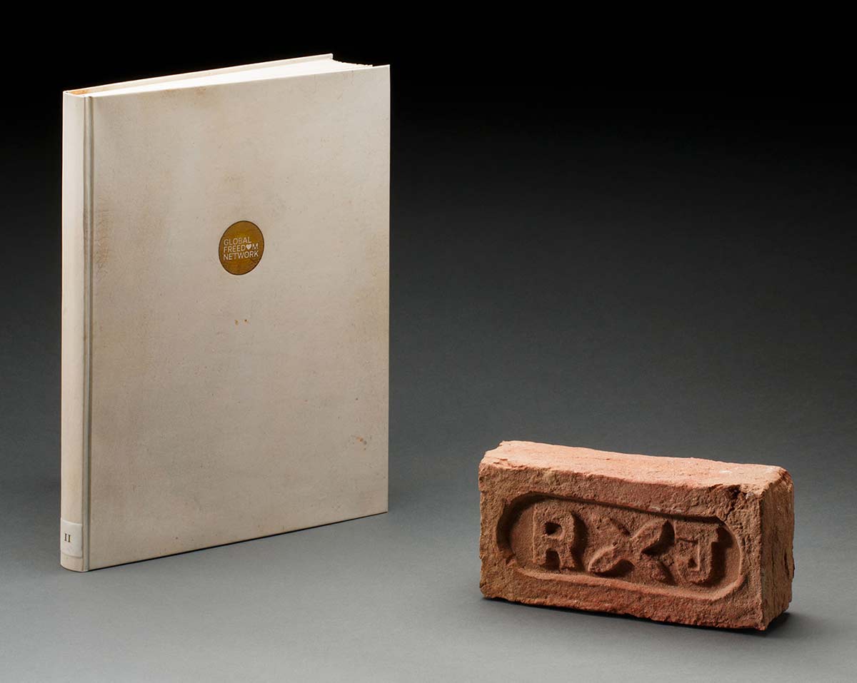 A cream coloured hard-covered book and a red-brown clay house brick marked with 'RXJ' on one face. The brick surface is uneven and friable.